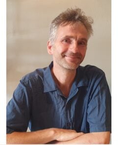 Portrait of a smiling man with short, graying hair wearing a blue collared shirt, sitting against a beige wall.