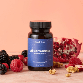Bottle of Akkermansia gut health supplement capsules surrounded by blackberries, raspberries, walnuts, and a halved pomegranate on a peach background.